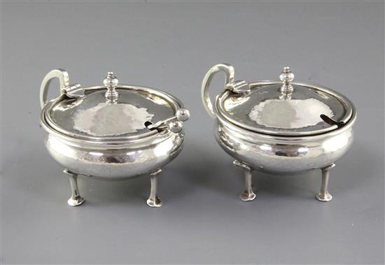 A pair of George V Arts & Crafts silver mustard pots with matching spoons, by Albert Edward Jones, width 90mm, weight 5.1oz/160grms.